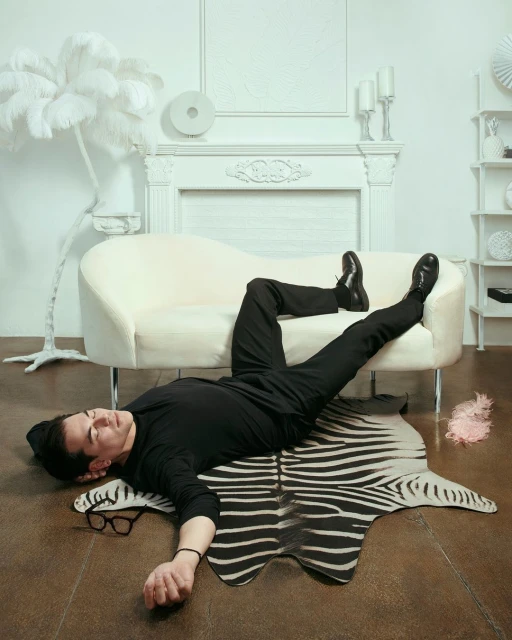 Man in black outfit lounging on a white sofa with a zebra print rug, glasses on the floor, in a minimalist white room.